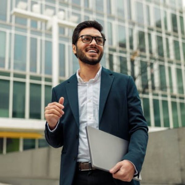 Smiling businessman in suit with laptop near modern office building is looking at side